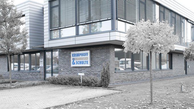Außenaufnahme der Krimphoff und Schulte Niederlassung in Rheine. Man sieht das Gebäude, an dem ein Schild mit dem Logo angebracht ist, sowie vor dem Gebäude einen Parkplatz und Grünflächen.